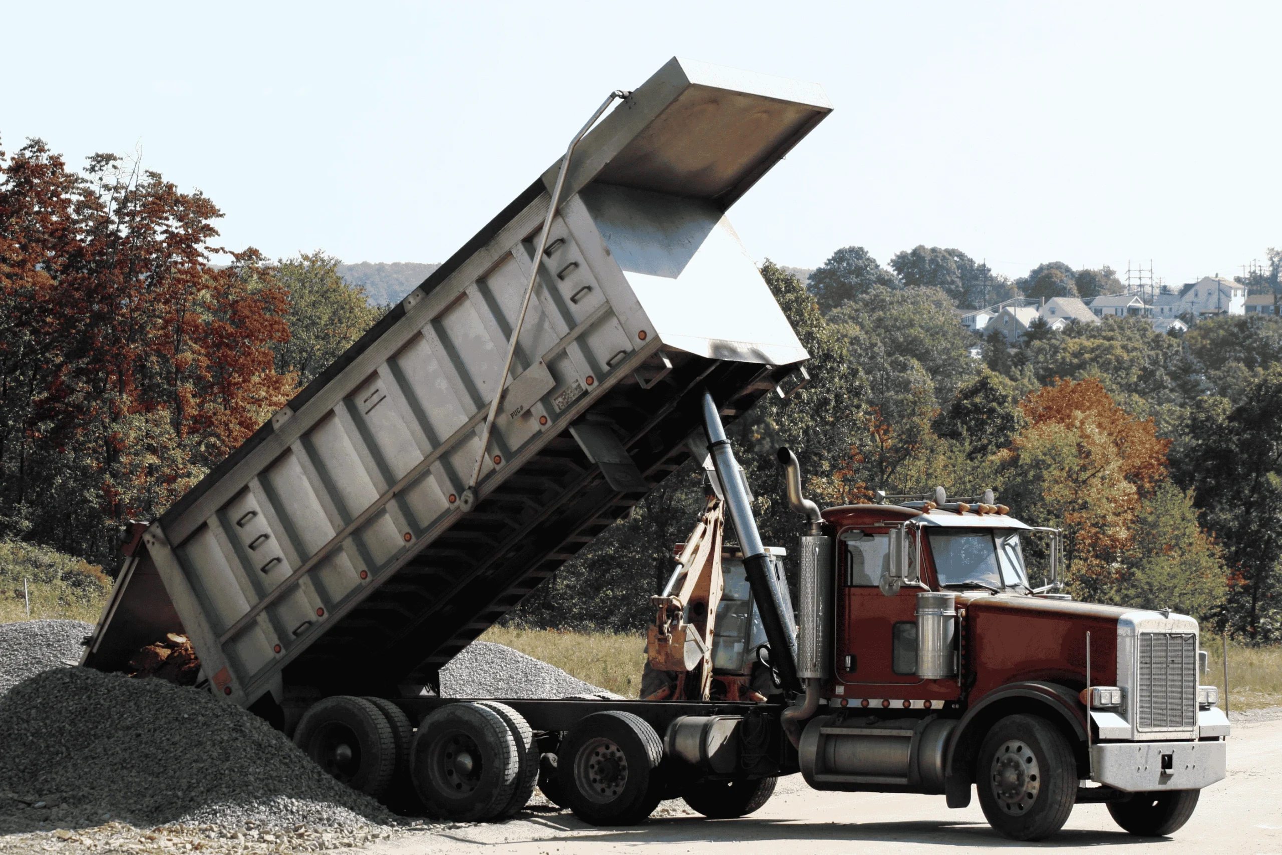 A photograph showing a red dump truck with its bed raised, actively unloading a pile of gravel at a rural construction site with a backhoe and autumn trees in the background, illustrating a scenario relevant to dump truck liability insurance.